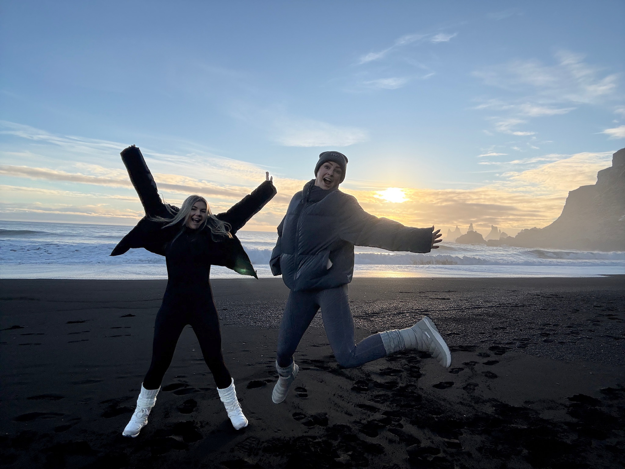Anastasia and Madison mid-jump on a black sand beach in Iceland at sunset.